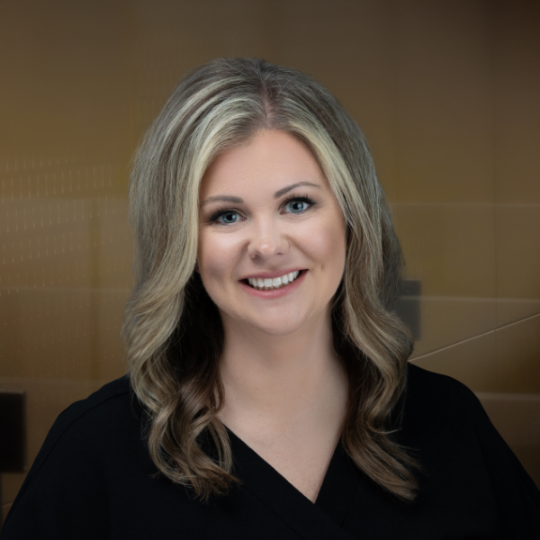 Smiling woman with blonde hair in a black V-neck top against a blurred brown background.